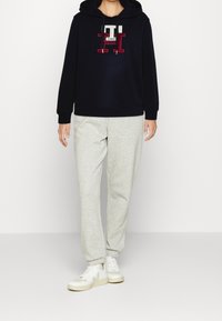 Felpa blu marina con logo, abbinata a pantaloni della tuta grigi chiari. L'outfit è completato da sneakers bianche, creando un look casual.