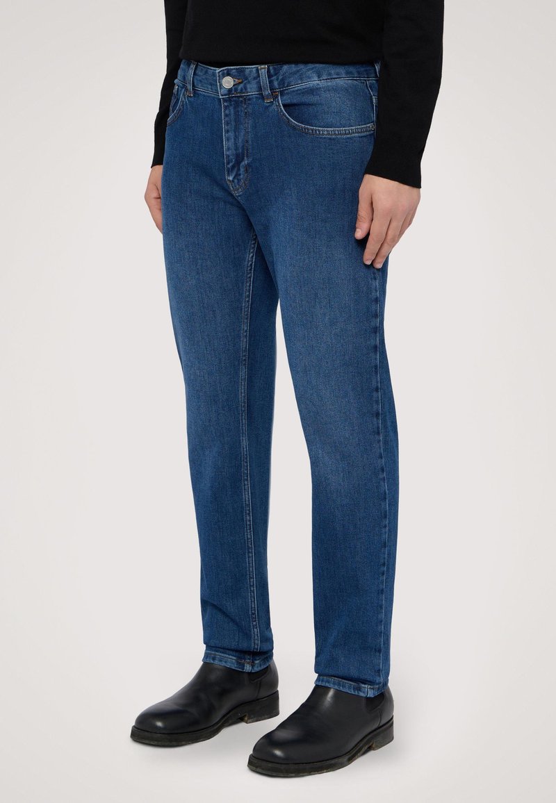 Vaqueros de denim azul de pierna recta con cinco bolsillos, costuras sutiles y cierre de botón metálico, combinados con botas negras de tobillo.