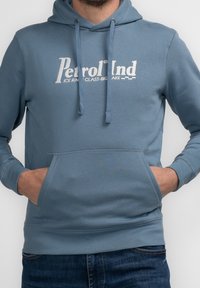 Modrá bavlněná mikina s kapucí, vybavená přední kapsou na klokaní způsob, stahovací kapucí a bílým logem na hrudi. Hladký materiál s pružnými manžetami.