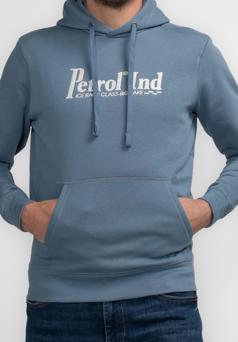 Modrá bavlněná mikina s kapucí, vybavená přední kapsou na klokaní způsob, stahovací kapucí a bílým logem na hrudi. Hladký materiál s pružnými manžetami.