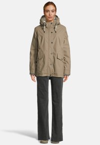Femme portant une veste à capuche beige avec des boutons et des poches à rabat, associée à un pantalon large gris foncé et des chaussures beiges.