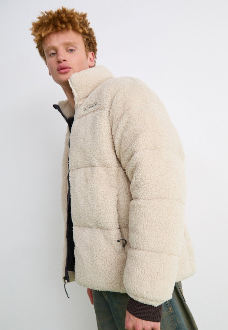 Cremefarbene, flauschige Puffjacke mit hohem Kragen, kontrastierenden Reißverschlüssen und elastischen Bündchen. Aus weichem, strukturiertem Fleece-Material.