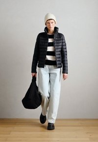 Melnā pūkainā jaka, baltmelnu svītru tops, gaiši zili bikses, nēsā melnu somu, adīta bēša cepure, melnas kurpes, iekštelpu vide.