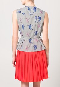 Top bez rukávů, světle šedý s potiskem ptáků a peplum pasem, spárovaný s plisovanou, jasně korálovou sukní. Tkanina vypadá hladce.