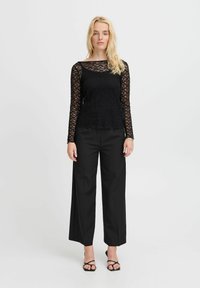 Schwarzes Spitzenoberteil mit langen Ärmeln und Rundhalsausschnitt, kombiniert mit hochtaillierten, schwarzen weiten Hosen. Das Model trägt schwarze Sandalen mit Riemchen.