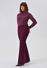 Uitlopende broeken in diep bordeauxrood, gecombineerd met een nauwsluitende, glinsterende paarse coltrui. Glad oppervlak, minimalistisch ontwerp met strakke lijnen.