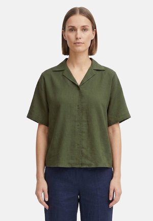 Donna con capelli castani lisci che indossa una camicia verde oliva a maniche corte con bottoni e pantaloni blu navy, in piedi davanti a uno sfondo semplice.