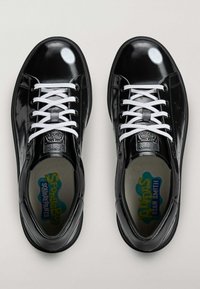 Crne tenisice od lakirane kože s bijelim vezicama, okruglim vrhom i logotipom Spužbob Skockani unutra. Imaju male perforacije sa strane.