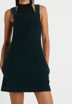 Kobieta w ciemnozielonej sukience sztruksowej na szerokich ramiączkach, na czarnym bezrękawnym topie, z rękami w kieszeniach sukienki.