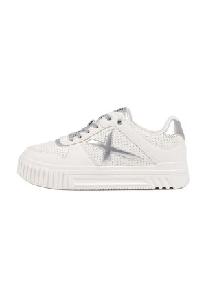 Zapatilla blanca de cuero perforado con detalles metálicos plateados. Presenta una suela gruesa de goma y cordones planos. Detalle de diseño en forma de X.