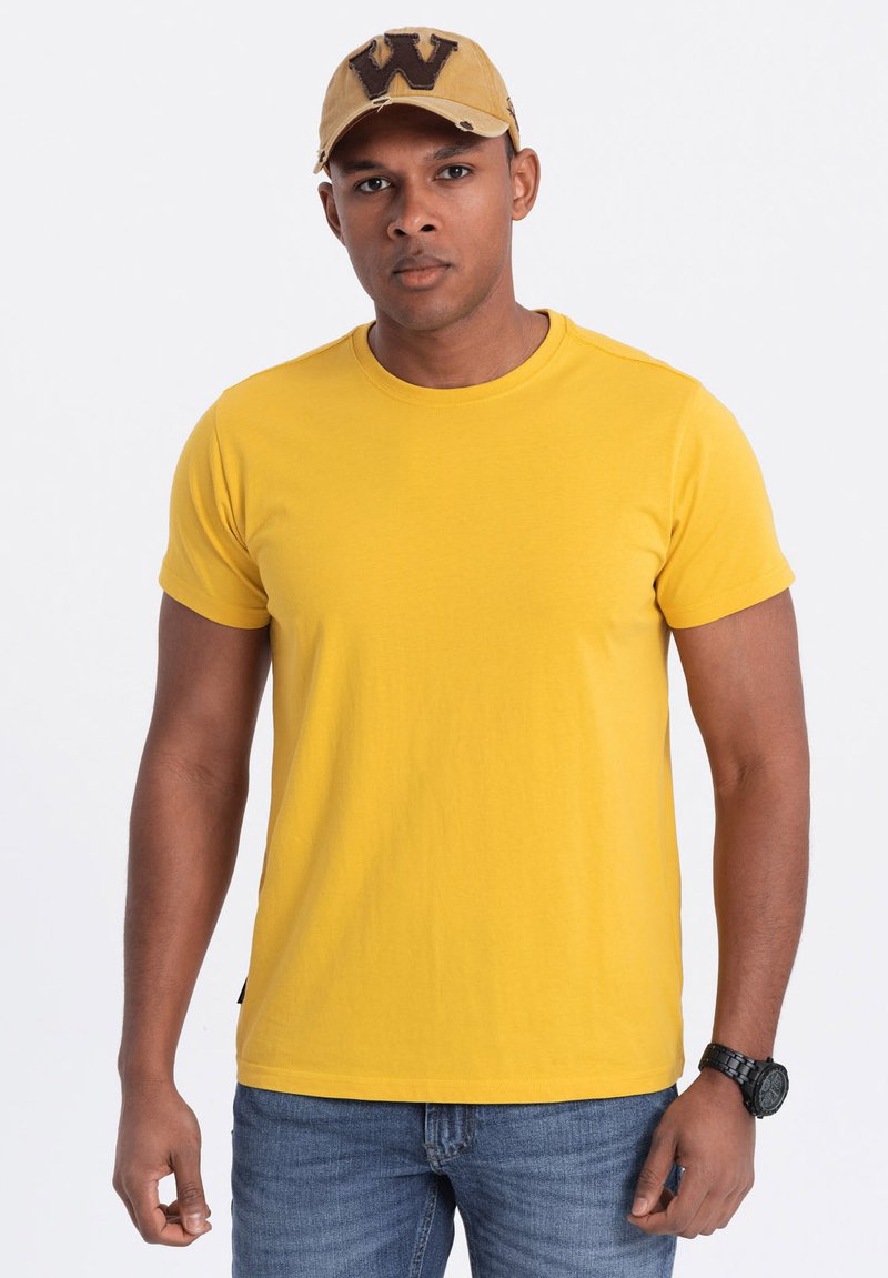 Gul bomulds-t-shirt med rund halsudskæring, korte ærmer og en afslappet pasform, parret med blå jeans og en beige kasket med et bogstav.