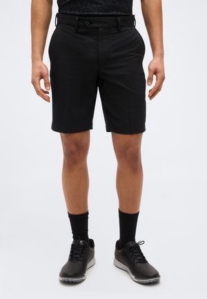 Hombre con pantalones cortos negros hasta la rodilla, calcetines negros hasta la mitad de la pantorrilla y zapatos deportivos negros, de pie contra un fondo blanco liso.