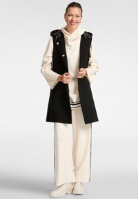 Frau, die lächelt, trägt einen schwarzen ärmellosen Mantel über einem cremegelfarbigen, weit geschnittenen Trainingsanzug mit schwarzen Streifen und beigen Knöchelschuhen.