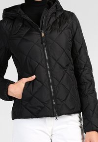 Veste matelassée noire avec fermeture éclair frontale, capuche et poches latérales ; présente un motif en losange et une texture lisse.