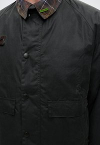 Μαύρο μπουφάν Barbour με καρό γιακά, κουμπιά με πατιλέτα, εμπρόσθιες τσέπες και μια μικρή πράσινη καρφίτσα SPY στο γιακά.