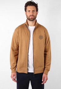Sweat à zip marron en tissu doux. Il présente un col haut, des poignets élastiques et un motif de flocon de neige brodé sur la poitrine.
