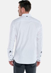 Mann in einem weißen Langarm-Hemd mit schwarzen Knopfdetails an den Manschetten, stehend mit dem Rücken zur Kamera, vor einem schlichten weißen Hintergrund.