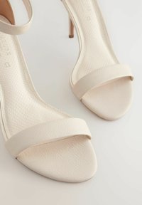 Sandalias de tacón beige con un upper de cuero sintético suave, diseño de tiras minimalistas y un tacón de aguja delgado. Detalle de textura sutil en la plantilla.