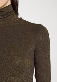 Brauner Rollkragenpullover aus strukturiertem Strickstoff mit funkelndem Finish. Enganliegender Schnitt mit umgeschlagenem Kragen, der einen subtilen Glanz zeigt.
