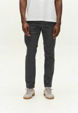 Person, die dunkelgraue Slim-Fit-Hosen, weiße Sneaker mit Gummisohlen und ein weißes T-Shirt vor einem einfarbigen hellen Hintergrund trägt.