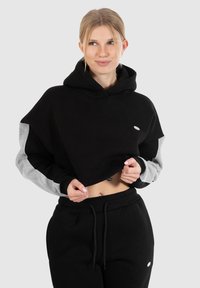 Zwart cropped hoodie met grijze mouwen, gemaakt van zachte stof. Beschikt over een capuchon met trekkoord en een klein logo op de borst. Gecombineerd met zwarte sweatpants.
