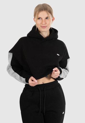 Schwarze, kurz geschnittene Kapuzenjacke mit grauen Ärmeln, aus weichem Material. Verfügt über eine Kordelzugkapuze und ein kleines Logo auf der Brust. Kombiniert mit schwarzen Jogginghosen.
