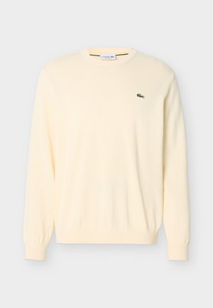 Krém színű hosszú ujjú Lacoste pulóver kerek nyakkivágással és apró zöld krokodil logóval a bal oldali mellrészen.