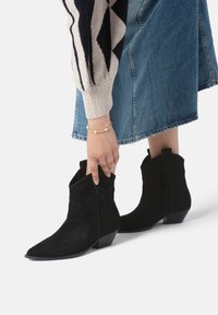 Botine negre din suede, cu vârf ascuțit, toc mic și pătrat, și cusături decorative. Purtați cu o fustă din denim și un pulover cu model.