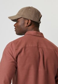 Casquette en velours côtelé brun et chemise à manches longues texturée couleur rouille avec un détail de bouton au col et un pli à l'arrière.