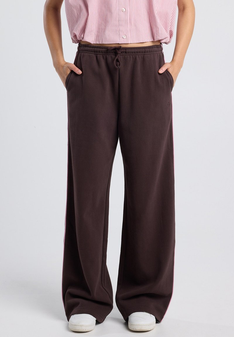 Person wearing loose brown sweatpants with pockets and white shoes, hands in pockets, against a plain light background.