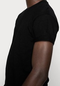 Czarny t-shirt z krótkim rękawem, wykonany z miękkiej, strukturalnej tkaniny; dopasowany krój z podwiniętymi mankietami, widok z boku.
