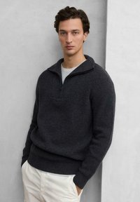 Ciemnoszary sweter z dzianiny z zamkiem przy kołnierzu, prążkowaną fakturą i klasycznym krojem, noszony na jasnoszarym T-shircie i zestawiony z jasnymi spodniami.