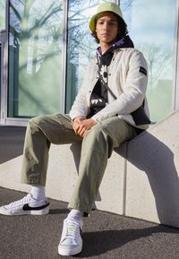 Witte pufferjas, zwarte bloemenhoodie, olijfgroene cargo-broek, witte sneakers met zwarte accenten en een lichtgroene bucket hat.