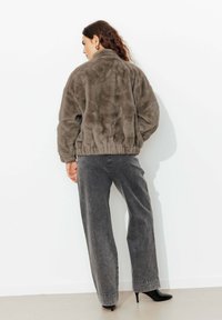 Veste bomber en fausse fourrure grise avec un col haut et un ourlet élastique. Associée à un pantalon large gris et des chaussures à talons noires.