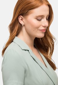 Orecchini a cerchio color oro con superficie texturizzata, indossati da una persona con lunghi capelli castano-rossicci, insieme a una blusa verde chiaro con rever.