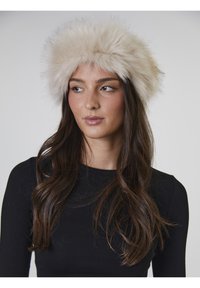 Gorra de falso pelo en color crema con una textura suave y esponjosa. Forma redonda con una banda ancha, llevada por un modelo con cabello oscuro y una blusa negra.
