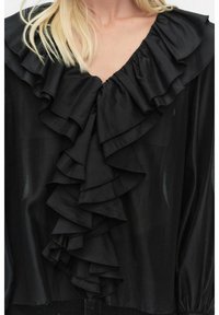 Svart blus med stor volangkrage, tillverkad av ett mjukt tyg. Har långa ärmar och en flödande design med strukturerade volanger.