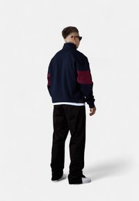 Veste bicolore marine et bordeaux avec col montant, associée à un pantalon noir et des baskets blanches, vue de l'arrière.