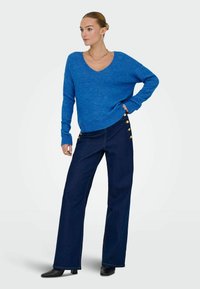 Femme portant un pull bleu à col en V, un pantalon large sombre avec des boutons dorés sur les côtés, des chaussures noires à talons et des bijoux dorés.