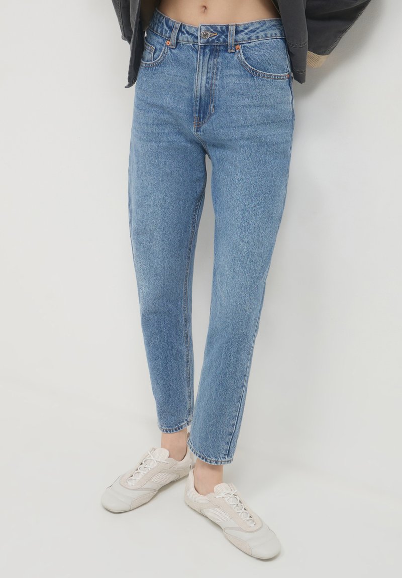 Jean taille haute bleu clair avec une coupe droite, dotés d'une fermeture à bouton et de cinq poches, associés à des baskets de couleur claire.