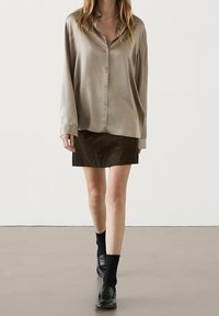Femme portant une blouse beige boutonnée, une mini-jupe marron foncé, des chaussettes noires et des mocassins noirs, debout devant un fond clair uni.
