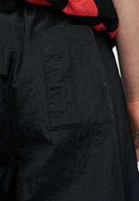 Κοντινό πλάνο υφασμάτινου μαύρου παντελονιού με υφή και πίσω τσέπη, σε συνδυασμό με πλεκτό ένδυμα σε κόκκινο και μαύρο και ορατό μπράτσο.