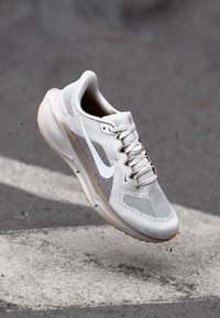 Bijela atletska cipela s mrežastim gornjim dijelom, sivi naglasci i bež potplat. Sadrži prepoznatljiv swoosh logo i vidljiv sustav vezanja.