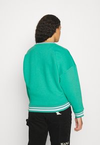 Tirkizna dukserica opuštenog kroja, s rebrastim ovratnikom i manžetama. Ima prugasti detalj na rubu. Nosena preko crnih ženskih hlača od kordura.