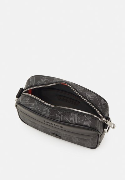 Trousse de toilette noire avec une finition texturée, présentant un motif géométrique, fermeture zippée et doublure intérieure rouge. Logo Lacoste sur le côté.
