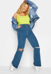 Jeans in denim blu a zampa d'elefante con strappi alle ginocchia, abbinati a una canotta gialla brillante e a una giacca di denim oversize azzurra.