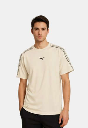 Młody mężczyzna w beżowej koszulce Puma z czarnym logo na środku i markowymi paskami na rękawach, stoi z jedną ręką w kieszeni.