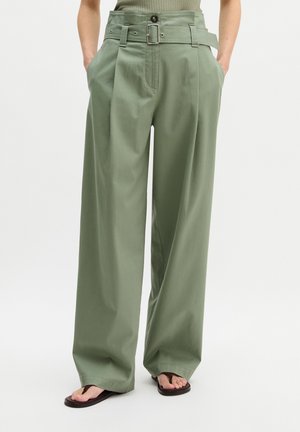 Vrouw draagt olijfgroene, wijde broek met hoge taille en ceintuur, handen in de zakken, gecombineerd met zwarte teenslippers tegen een witte achtergrond.