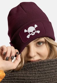 Maroon beanie hat made of ribbed knit fabric with a large white skull and crossbones patch on the front. Warm and fitted design.