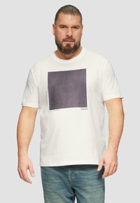 Witte katoenen t-shirt met een donkere vierkante grafische print op de voorkant, ronde halslijn en korte mouwen. In combinatie met lichtblauwe jeans.